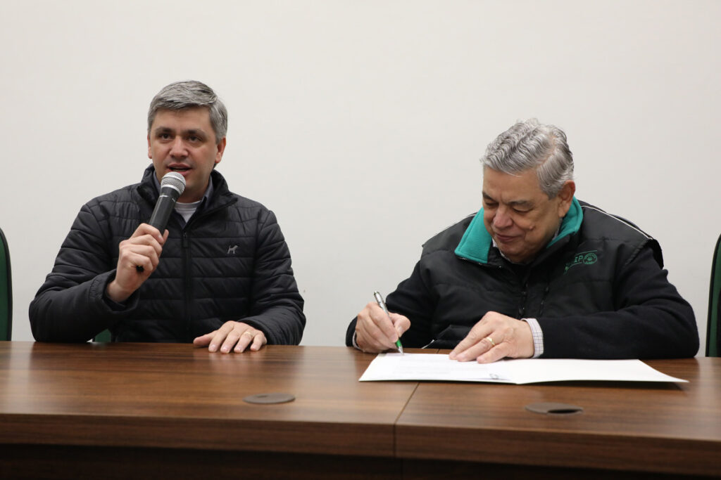 Dois homens de cabelo grisalho sentados à mesa. O primeiro, à esquerda, está falando em um microfone, o segundo, à direita, está assinando um documento.