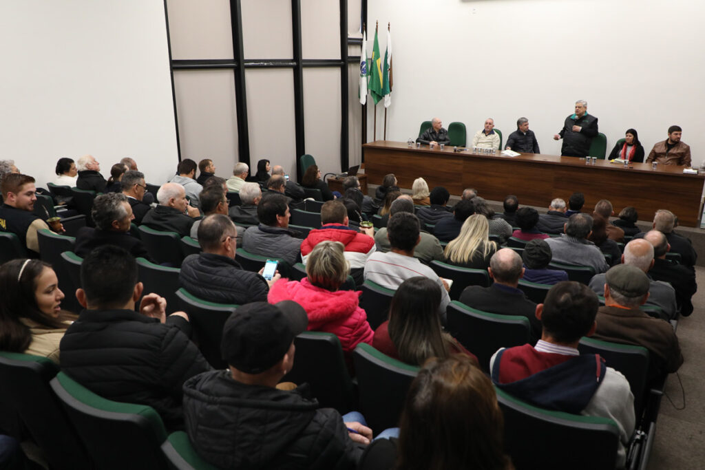 Auditório da Fetaep lotado assiste à cerimônia de entrega dos equipamentos. Na frente do público, há uma mesa de madeira com presença de seis pessoas, uma delas está em pé discursando em um microfone. À esquerda da mesa, estão as bandeiras do Paraná, do Brasil e de Curitiba.