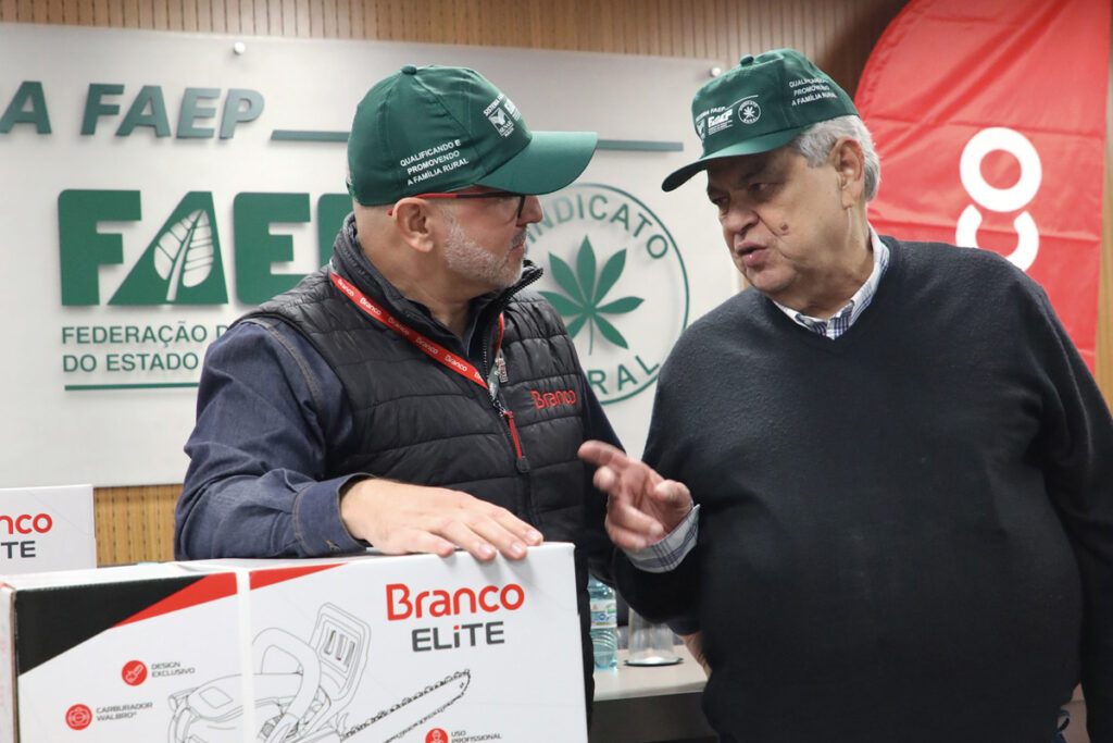 Dois homens usando o boné verde do Sistema FAEP/SENAR-PR conversam. O primeiro homem se apoia sobre uma caixa branca, com o desenho de uma motosserra e detalhes em vermelho e preto, da Branco Motores.