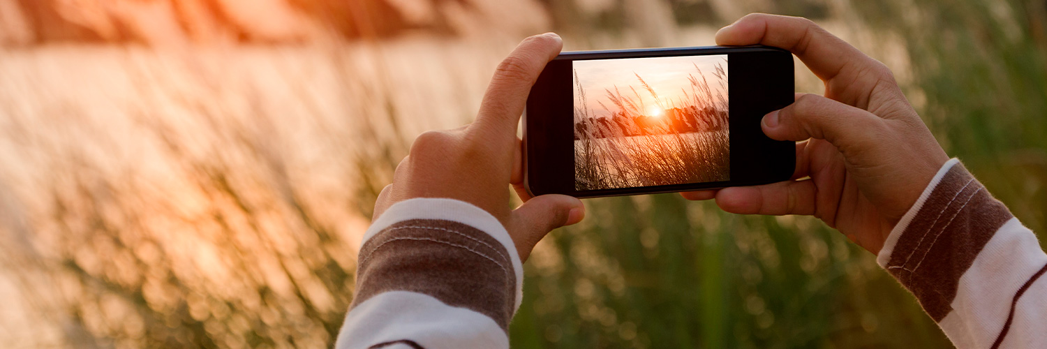 Pessoa tirando foto de uma paisagem com pôr do sol com um celular na horizontal.