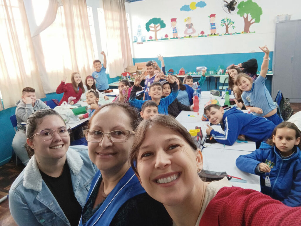 Professoras e alunos sorriem para foto tirada dentro de sala de aula lotada de crianças