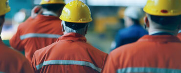 Bombeiros uniformizados com capacetes de costas para a câmera, prestando atenção na explicação de alguém que não aparece na imagem