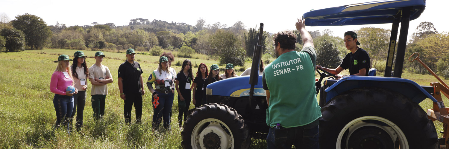 Alunos de colégio agrícola em aula a campo com trator em primeiro plano e grupo de estudantes ao fundo, em área com gramado